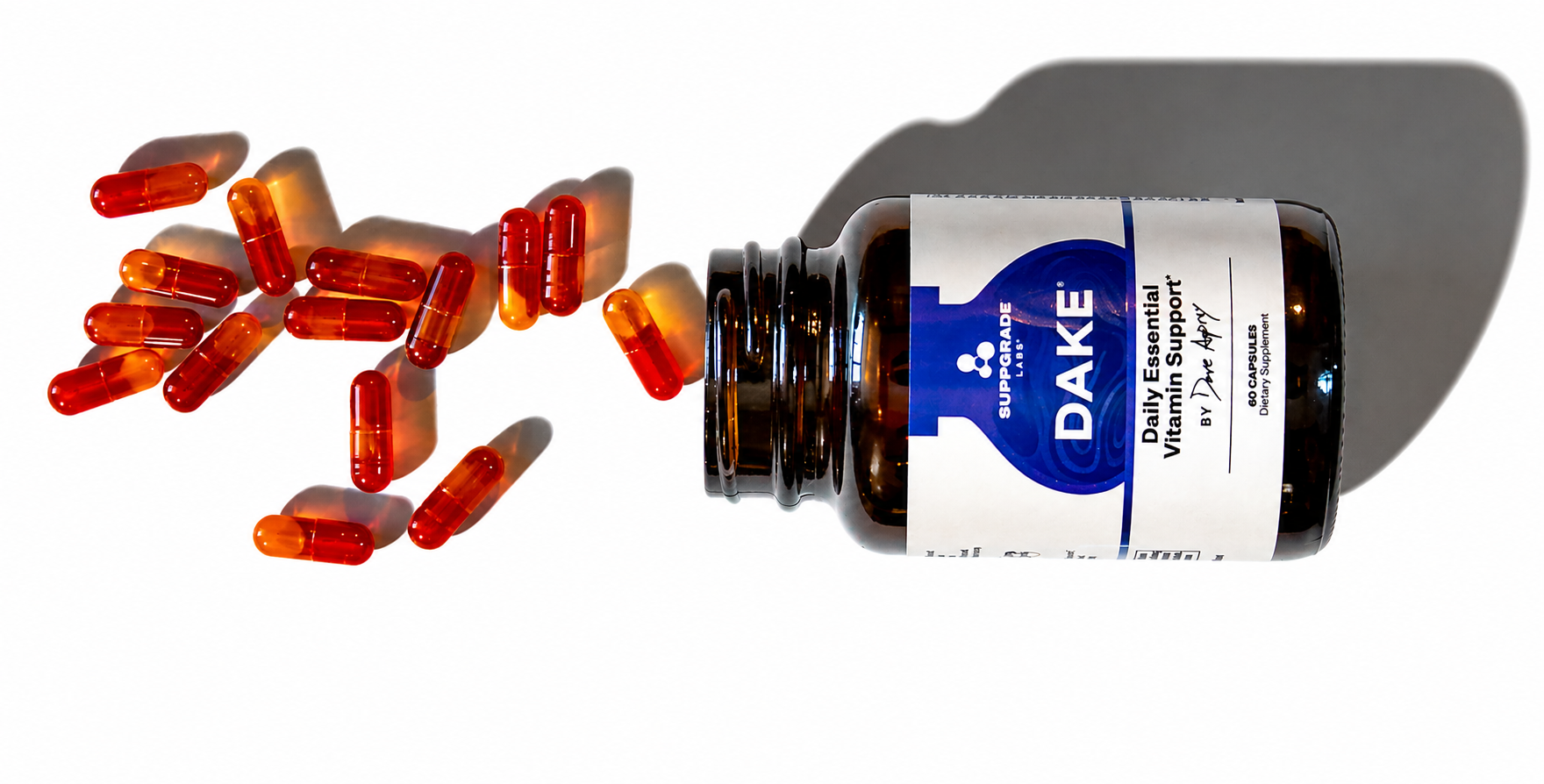 Amber bottle of DAKE daily essential vitamin capsules with orange and red capsules spilling out on a white background.