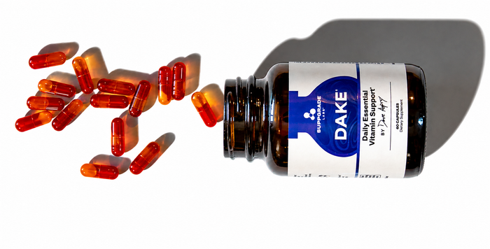 Amber bottle of DAKE daily essential vitamin capsules with orange and red capsules spilling out on a white background.