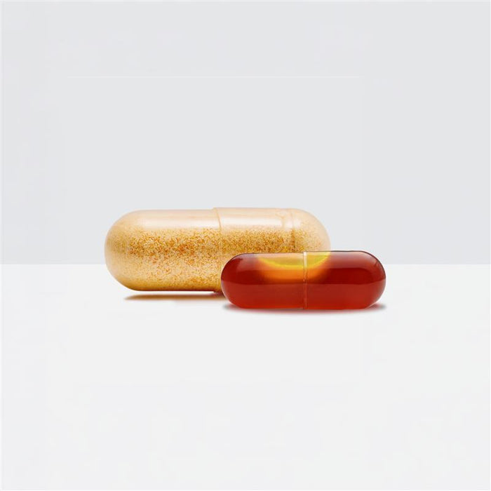 Two supplement capsules side by side — a large golden powder-filled capsule (old formulation) behind a smaller, deep amber softgel capsule (new formulation), photographed on a clean white surface.