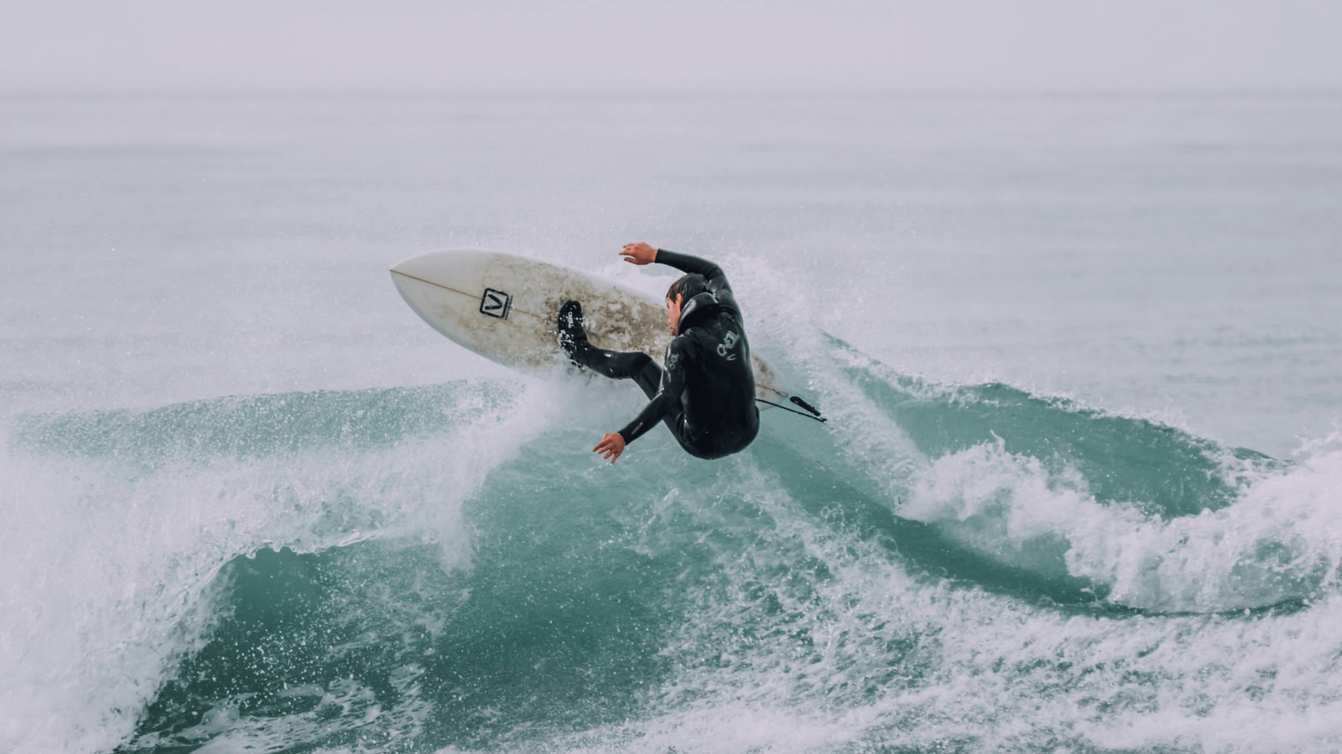 Surfer in a black wetsuit performing an aerial maneuver on a wave with turbulent water spray in the ocean.