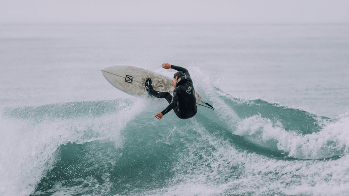 Surfer in a black wetsuit performing an aerial maneuver on a wave with turbulent water spray in the ocean.