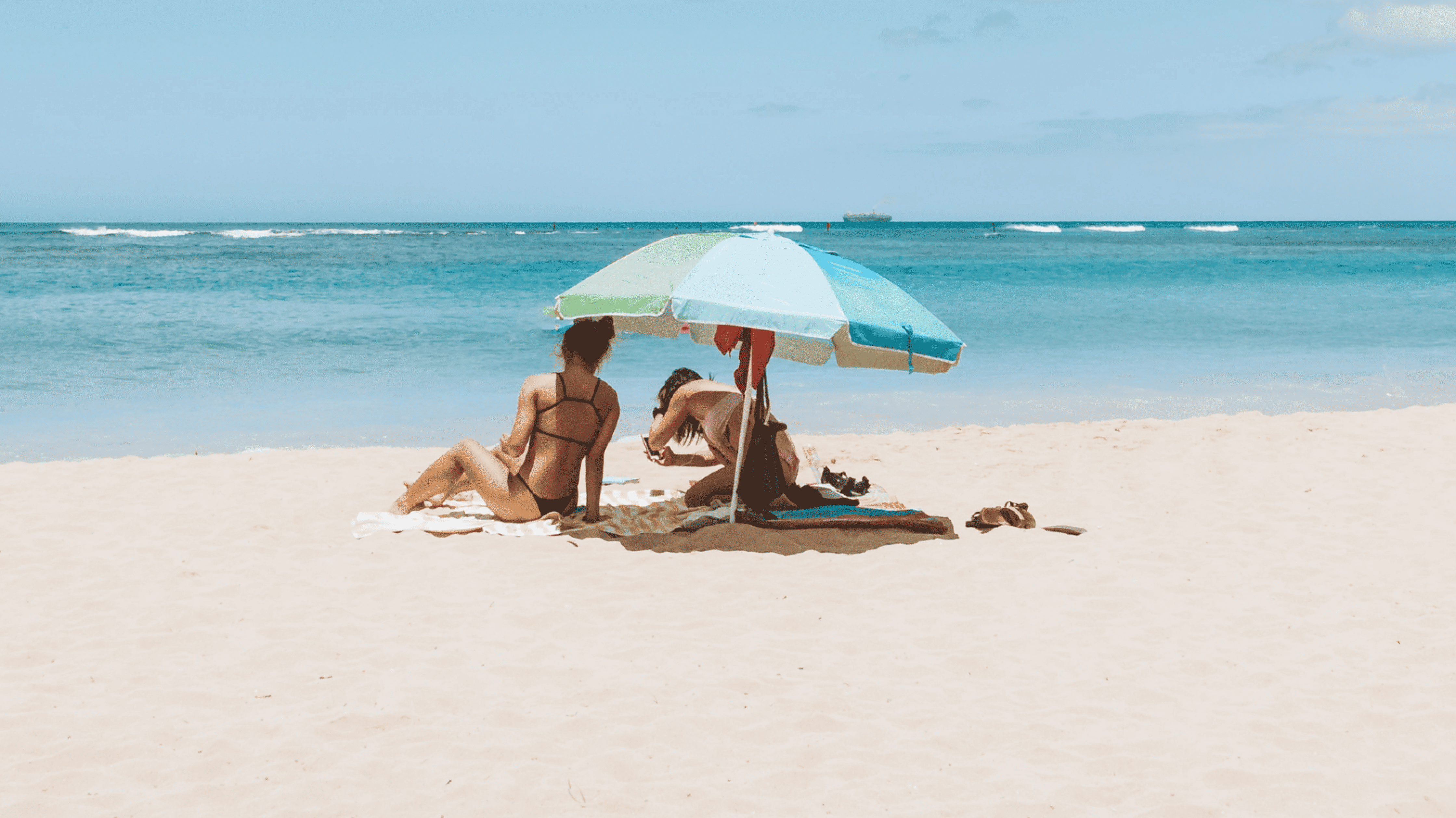 Two women in bikinis relax under a blue and green beach umbrella on sandy shore with ocean waves and clear sky.