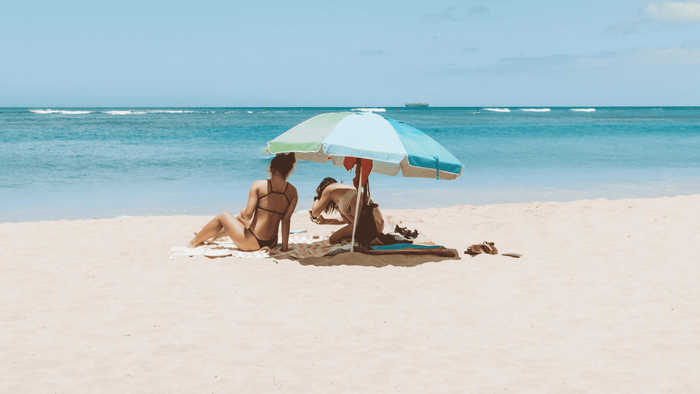 Two women in bikinis relax under a blue and green beach umbrella on sandy shore with ocean waves and clear sky.