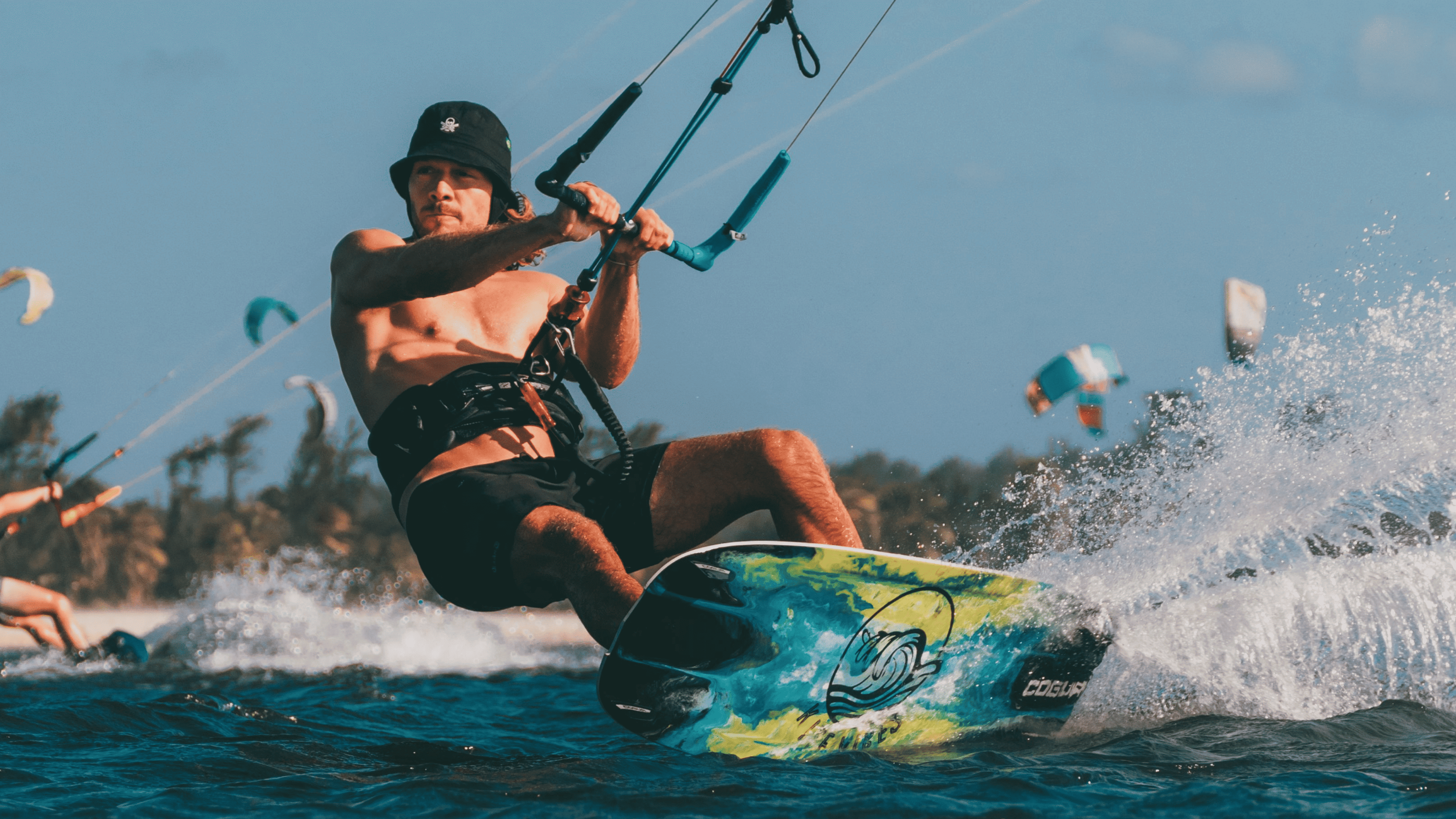 Man wearing a black hat kiteboarding on the water, creating a spray with colorful kites and trees in the background.