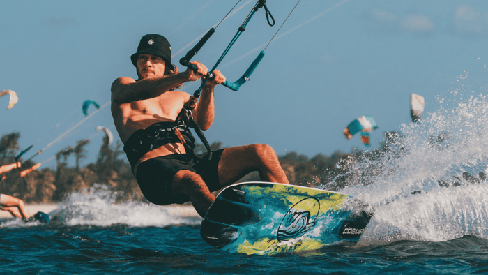 Man wearing a black hat kiteboarding on the water, creating a spray with colorful kites and trees in the background.