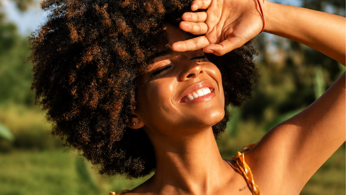 Smiling woman with curly hair shielding her eyes from sunlight, standing outdoors with a blurred green background.