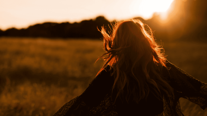 Woman with long hair stands in a sunlit field at sunset, facing away with arms slightly extended, bathed in warm golden light.