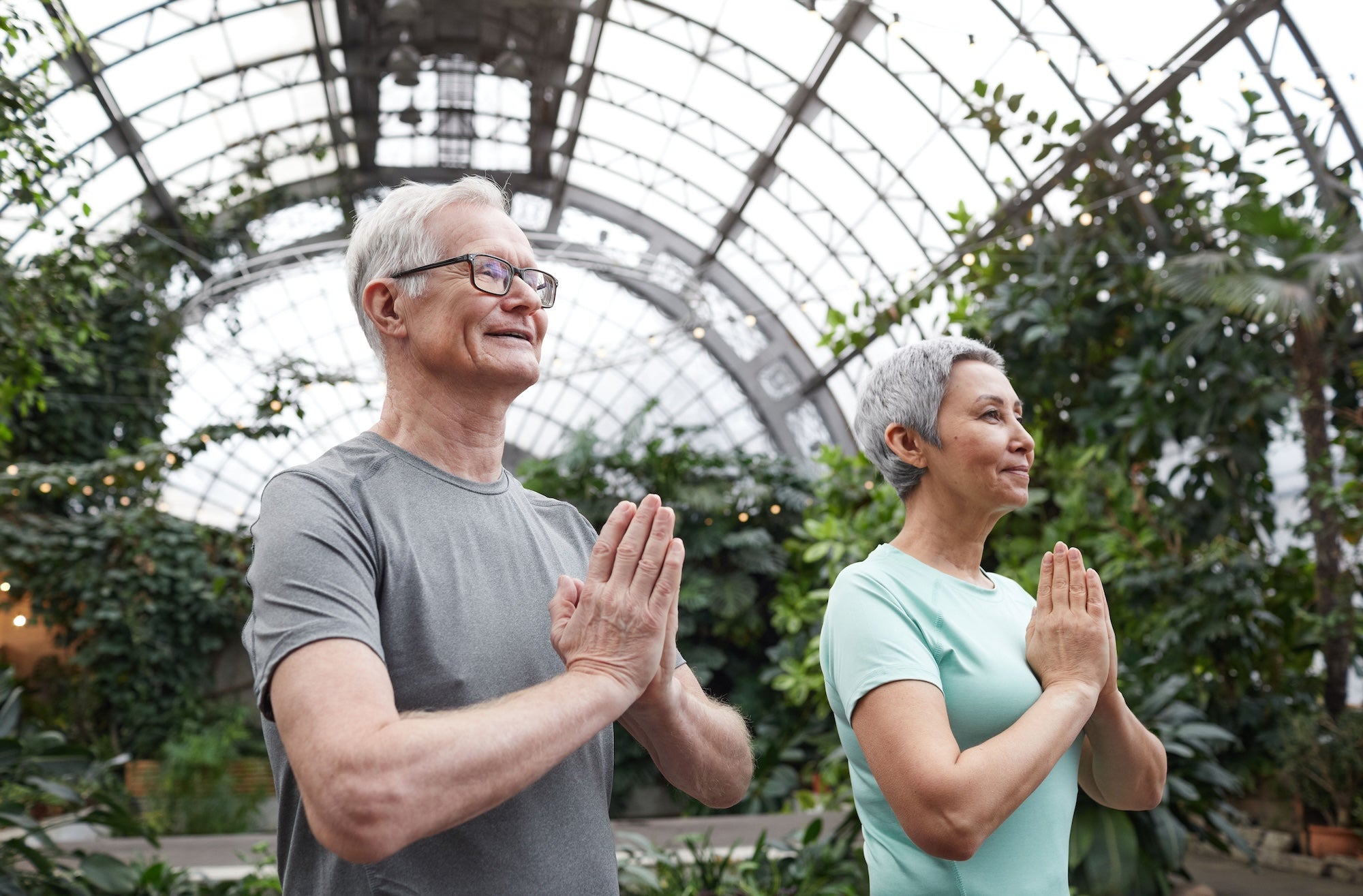 Two older adults practicing yoga with hands in prayer pose inside a lush greenhouse with a glass ceiling.