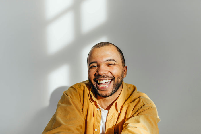 A man laughing warmly, wearing a mustard yellow button-up shirt over a white t-shirt, against a light gray wall with soft window light and shadows in the background.